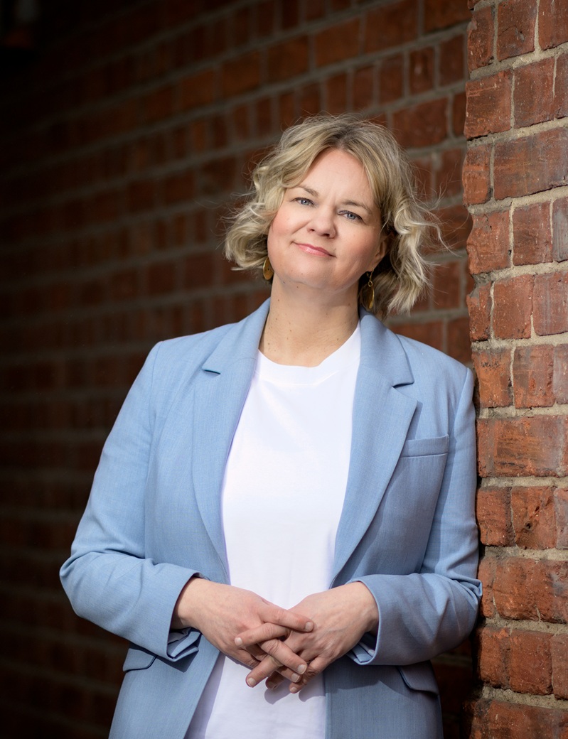 Toimitusjohtaja Tiina Ristikari nojaa hymyillen tiiliseinään. Hänellä on yllään valkoinen paita ja vaaleansininen jakku.